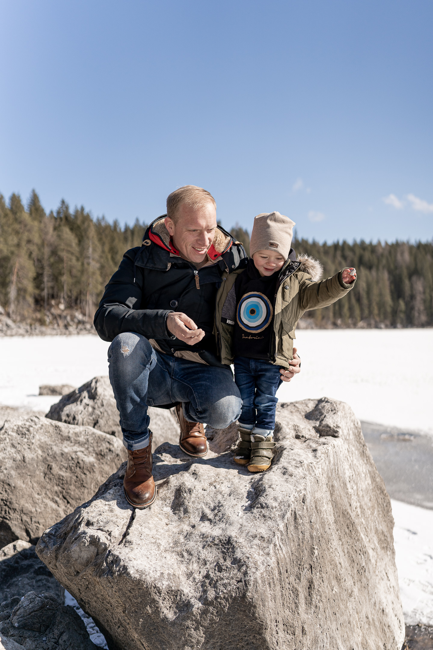 Junge sitzt mit Vater auf einem Felsen und schmeißen Steine auf dem zugefrorenen Eibsee im Winter während eines Familienshootings